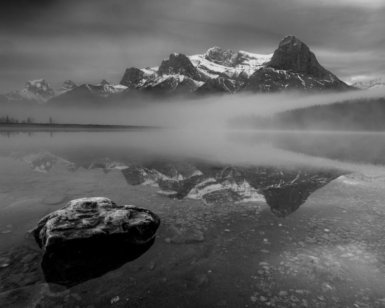 A black and white image of a rock in a lake in front of a mountain reflection.